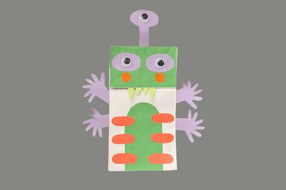 A martian puppet made from a silver paper bag with a green face and stomach with red stripes, purple eyes and arms and four large fangs. 