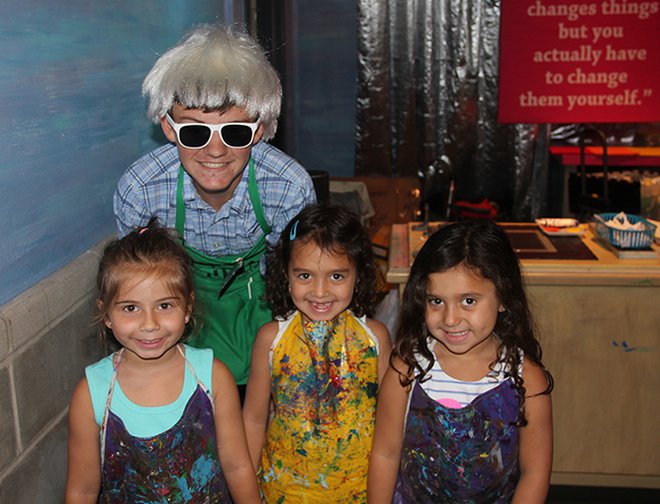 A Museum volunteer wearing a Andy Warhol wig standing with children. 
