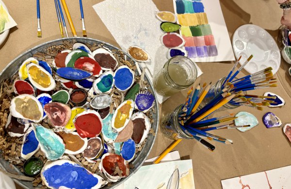 A mix of painted oyster shells, paintbrushes in glass jars, and paint palettes on a table covered with butcher paper. 