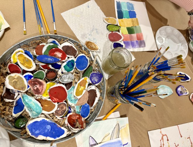 A mix of painted oyster shells, paintbrushes in glass jars, and paint palettes on a table covered with butcher paper. 
