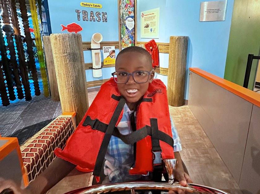 Boy wearing orange life vist at the wheel of a fishing boat.  Pilings are visible behind him.