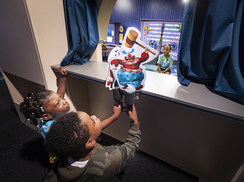 Two children holding puppets over a small stage while another child watches from the audience. 