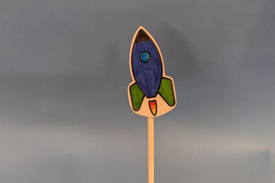 A small drawn blue and green rocketship attached to a wooden dowel. 