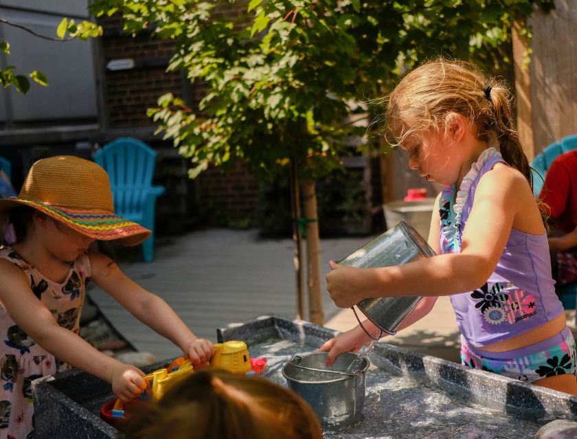 Two children wearing bathing suits in the H2Oh! area playing at the water table with metal buckets and plastic boats. 