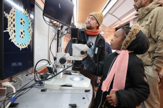 Girl in jacket and scarf looks up at screen connected to microscope. 