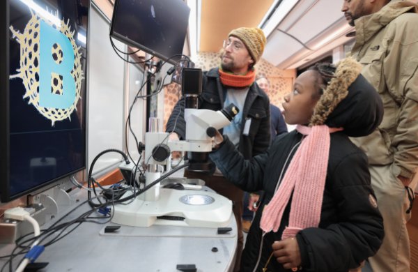 Girl in jacket and scarf looks up at screen connected to microscope. 