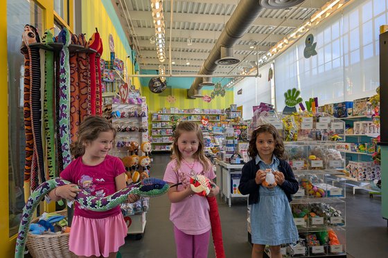 Three children standing in the Museum Store looking at the camera smiling while holding a stuffed animal. 