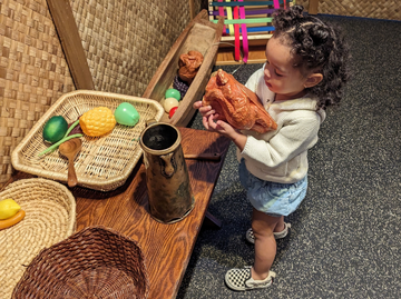 A child standing at a table in a straw house holding a plastic cooked chicken. 