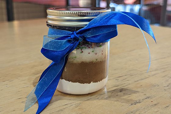 A glass jar with a blue bow on it filled with cocoa, sugar, and sprinkles.