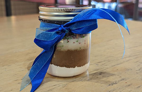 A glass jar with a blue bow on it filled with cocoa, sugar, and sprinkles.