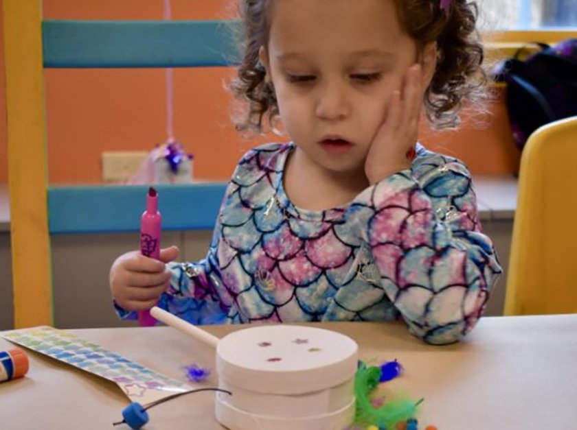 A child sitting at a table with her hand on their cheek while coloring a paper drum.