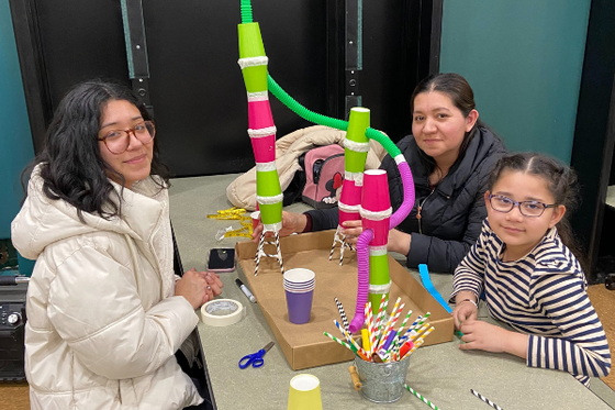 A child and two adults sitting at a table with their rollercoaster made from cups, straws and tubes. 