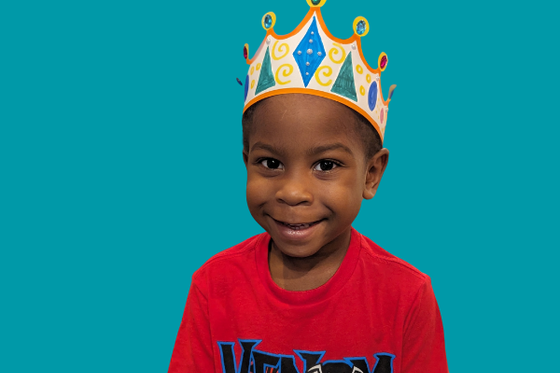 A child looking at the camera while wearing a decoated paper crown. 