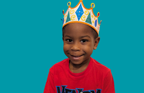 A child looking at the camera while wearing a decoated paper crown. 