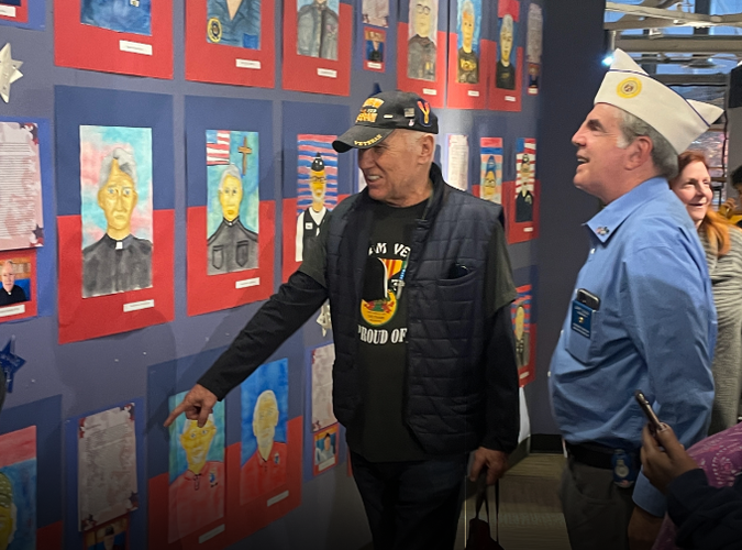 Two men wearing armed forces clothes while looking at a gallery of portaits of veterans of the armed forces. 