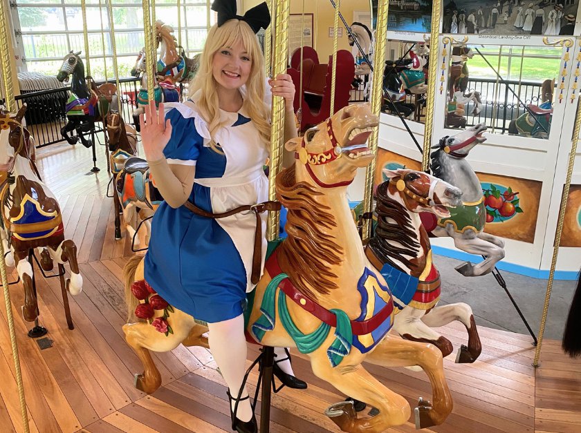 A woman dressed in an Alice in Wonderland costume is waving while sitting on a white carousel horse.