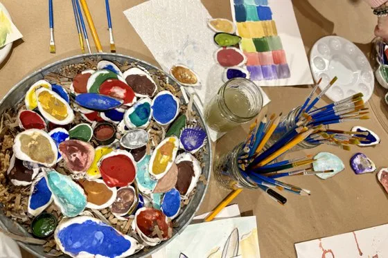 A mix of painted oyster shells, paintbrushes in glass jars, and paint palettes on a table covered with butcher paper.