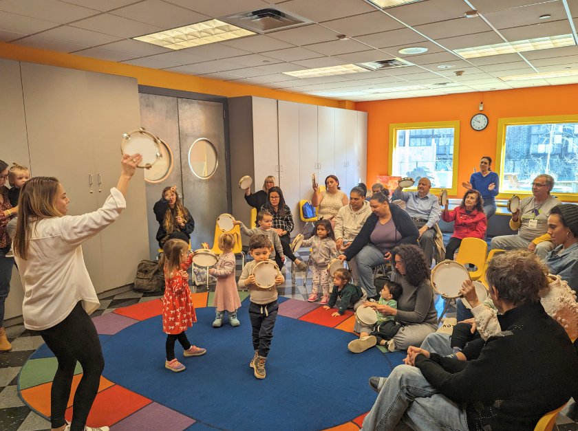 A group of children dancing on a carpet while holding instruments with adults sitting around the perimeter in chairs.