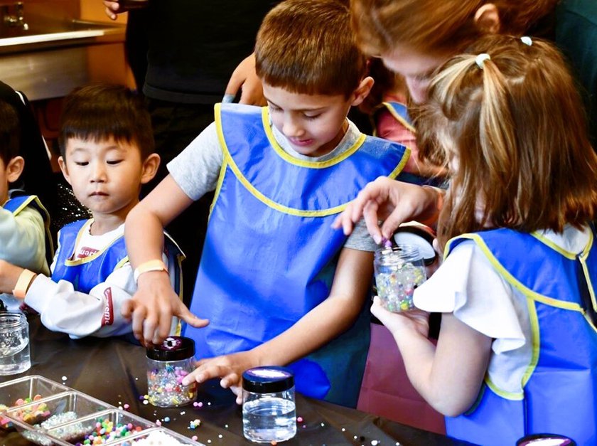 A group of children standing at a table wearing aprons while adding colorful beads and pom poms to their slime.