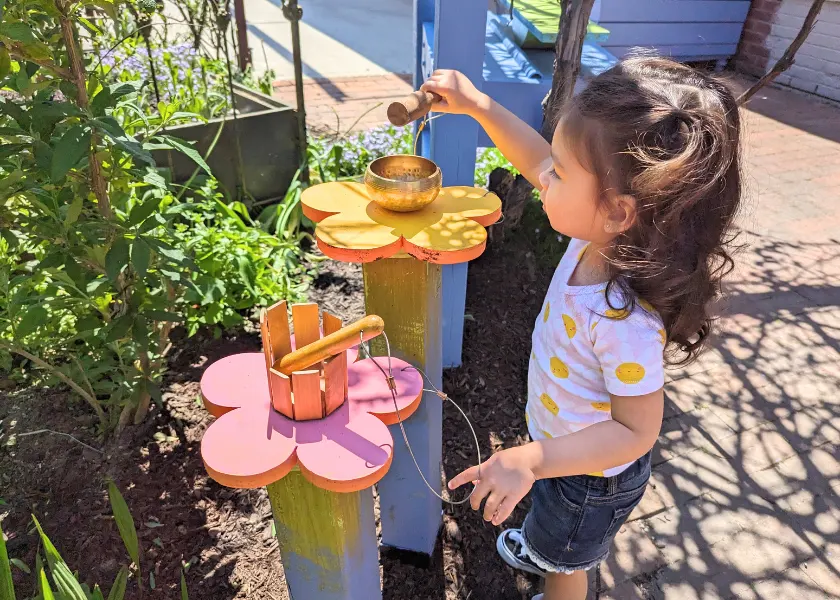 A child standing outside holding a drum mallet while playing a small metal drum.