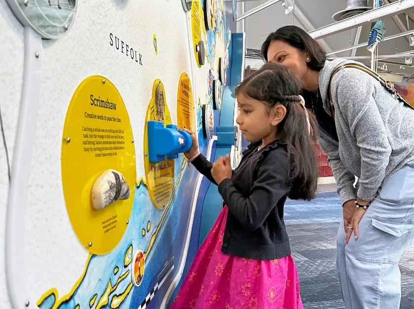 Adult and child stand before map featuring a piece of scrimshaw and a whale oil sniff activity.