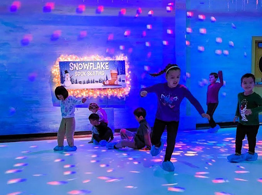 A group of kids wearing covers over their shoes while 'skating' on synthetic ice with signage behind them reading Snowflake Sock Skating.