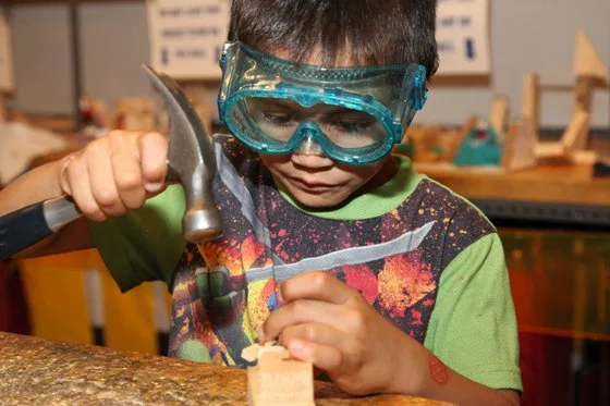 Boy using a hammer and nail to create wooden project while wearing safety goggles.