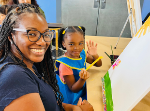 A staff member smiles with a child at an art easel.