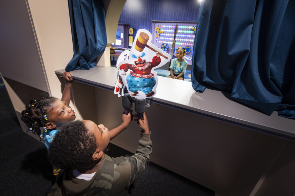 Two children holding puppets over a small stage while another child watches from the audience.