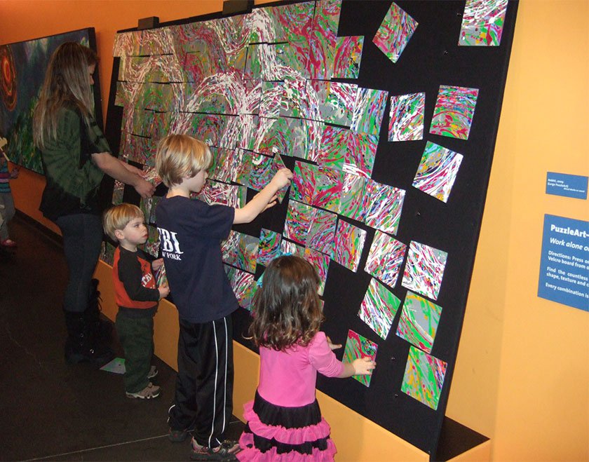 Children and an adult moving square pieces of painted canvas in order to connect white lines throughout painting.