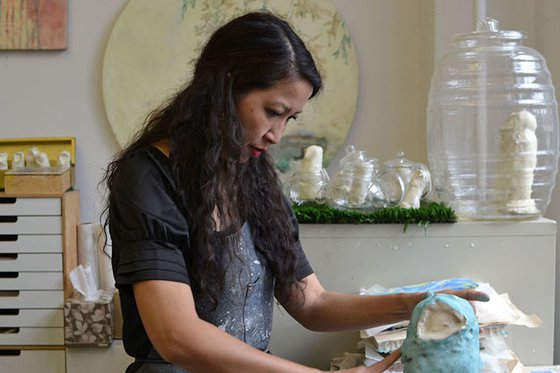 Artist in her studio creating sculpture with various pieces of artwork on the wall in the background.