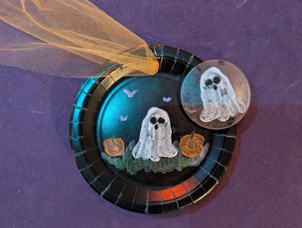 A black paper plate decorated with a painted ghost, pumpkins and bats, and orange tule.