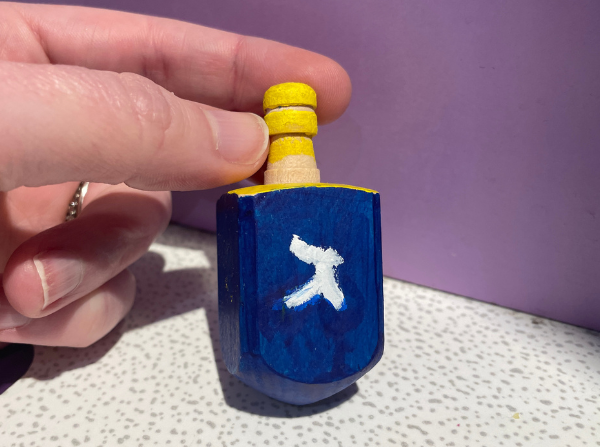 Hand holding a dreidel painted blue with yellow top. The third letter of the Hebrew Aleph-Bet, Gimmel (ג) is painted in white