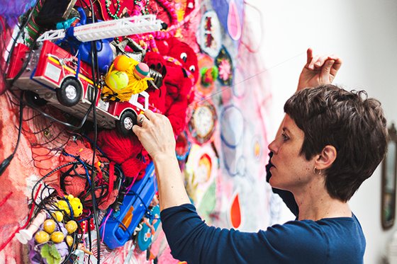 Artist with sewing needle adding brightly colored fabric and discarded toys to a canvas.