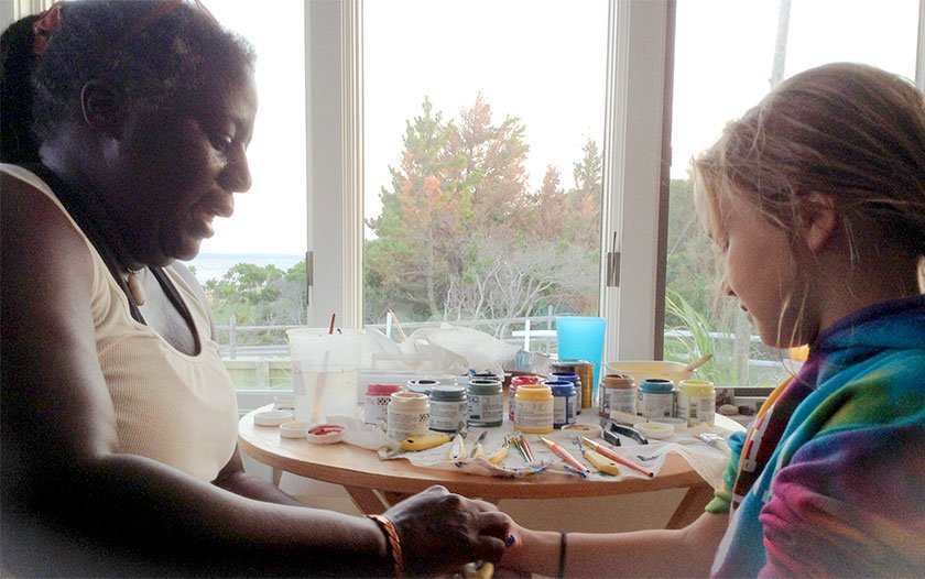 Artist Kooy sitting and speaking with young artist painting her hand.