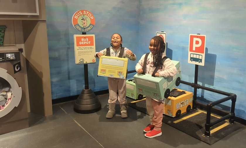Two children standing in the Mo Willems exhibit wearing cardboard cars over their waist.